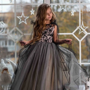 May include: A young girl wearing a black and pink tulle dress with floral embroidery. The dress has a cape and is worn with a tiara. The girl is standing in front of a window with string lights in the shape of stars.