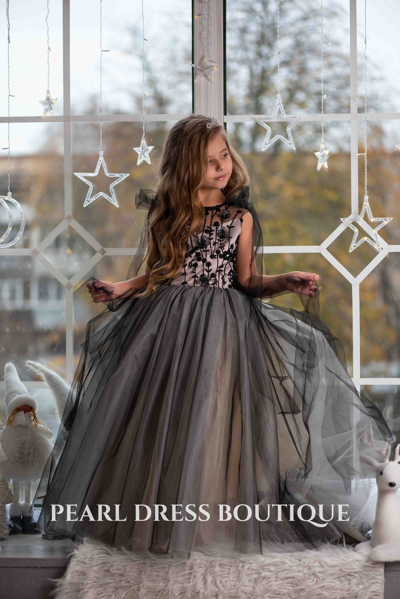 May include: A young girl wearing a black and pink tulle dress with floral embroidery. The dress has a cape and is worn with a tiara. The girl is standing in front of a window with string lights in the shape of stars.