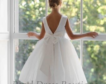 Puede incluir: Un vestido de tul blanco con un corpiño de satén y un gran lazo en la espalda. El vestido tiene la espalda baja y está decorado con detalles de perlas. El vestido lo lleva una niña de pie frente a una ventana.