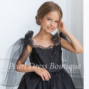 Puede incluir: Una ni&ntilde;a peque&ntilde;a con un vestido de tul negro con detalles de encaje y mangas transparentes negras. El vestido tiene un escote de coraz&oacute;n y una falda completa. La ni&ntilde;a sonr&iacute;e y mira a la c&aacute;mara. El texto "Pearl Dress Boutique" es visible en la imagen.
