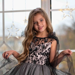 May include: A young girl wearing a black and pink tulle dress with floral embroidery and a black tulle cape. She is wearing a small tiara and smiling at the camera. The text "PEARL DRESS BOUTIQUE" is visible at the bottom of the image.