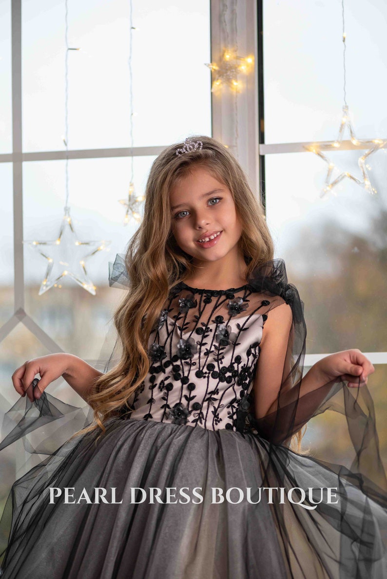 May include: A young girl wearing a black and pink tulle dress with floral embroidery and a black tulle cape. She is wearing a small tiara and smiling at the camera. The text "PEARL DRESS BOUTIQUE" is visible at the bottom of the image.