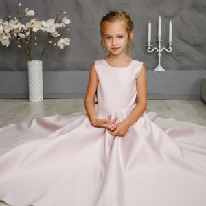 Pu&ograve; includere: Una bambina che indossa un abito di raso rosa chiaro con una gonna ampia. &Egrave; seduta su un pavimento di legno chiaro con un vaso di fiori bianco e un candelabro bianco dietro di lei.