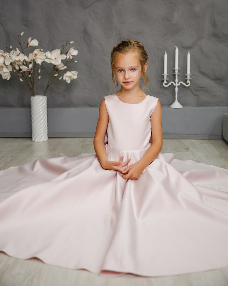 Pu&ograve; includere: Una bambina che indossa un abito di raso rosa chiaro con una gonna ampia. &Egrave; seduta su un pavimento di legno chiaro con un vaso di fiori bianco e un candelabro bianco dietro di lei.