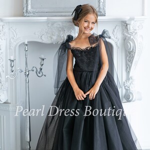 Puede incluir: Un vestido de tul negro con un corpi&ntilde;o de encaje y grandes lazos en los hombros. El vestido es de largo hasta el suelo y tiene una falda completa. La modelo lleva un lazo negro en el pelo. "Pearl Dress Boutique" est&aacute; escrito en la parte inferior de la imagen.