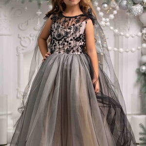 May include: A young girl wearing a black and gray tulle dress with a black floral design and a black cape. The dress has a fitted bodice and a full skirt. The girl is standing in front of a white fireplace with a silver garland and ornaments.