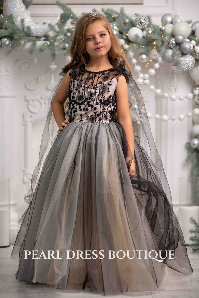 May include: A young girl wearing a black and gray tulle dress with a black floral design and a black cape. The dress has a fitted bodice and a full skirt. The girl is standing in front of a white fireplace with a silver garland and ornaments.