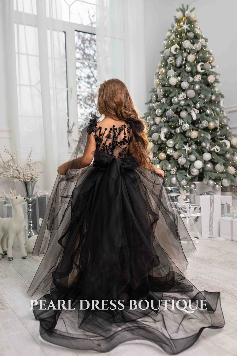May include: A young girl wearing a black tulle dress with a beaded bodice and a large bow in the back. The dress has a long train and is perfect for a special occasion. The girl is standing in front of a Christmas tree.