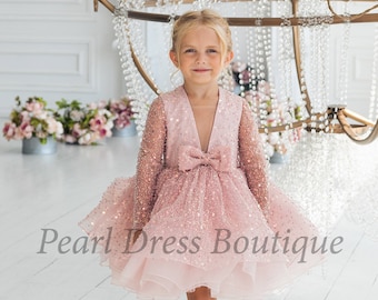 May include: A young girl wearing a pink sparkly dress with a bow at the neckline. The dress has long sleeves and a layered skirt. The girl is standing in front of a white background with a chandelier in the background. The text "Pearl Dress Boutique" is visible at the bottom of the image.