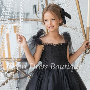 Puede incluir: Una ni&ntilde;a peque&ntilde;a con un vestido de tul negro con detalles de encaje y un lazo negro en el pelo. El vestido tiene mangas transparentes con lazos en los hombros. La ni&ntilde;a est&aacute; de pie frente a una l&aacute;mpara de ara&ntilde;a de cristal.