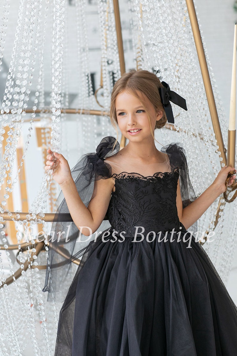 Puede incluir: Una ni&ntilde;a peque&ntilde;a con un vestido de tul negro con detalles de encaje y un lazo negro en el pelo. El vestido tiene mangas transparentes con lazos en los hombros. La ni&ntilde;a est&aacute; de pie frente a una l&aacute;mpara de ara&ntilde;a de cristal.