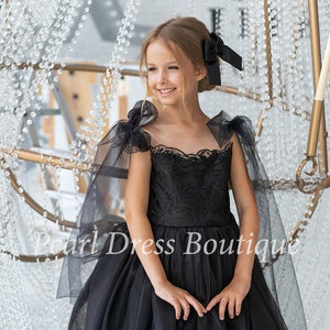 Puede incluir: Una ni&ntilde;a peque&ntilde;a lleva un vestido de tul negro con un corpi&ntilde;o de encaje y mangas transparentes negras. El vestido tiene una falda completa y est&aacute; decorado con bordados negros. La ni&ntilde;a sonr&iacute;e y mira hacia un lado. El vestido es una opci&oacute;n hermosa y elegante para una ocasi&oacute;n especial.