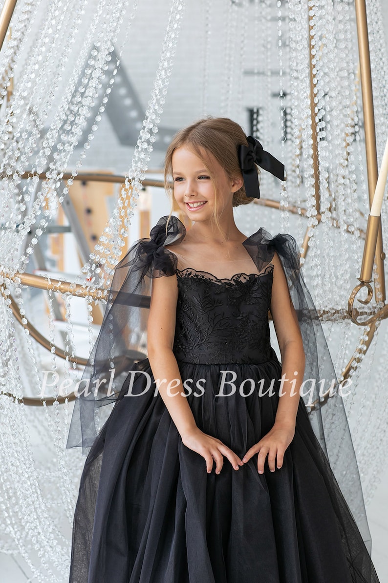 Puede incluir: Una ni&ntilde;a peque&ntilde;a lleva un vestido de tul negro con un corpi&ntilde;o de encaje y mangas transparentes negras. El vestido tiene una falda completa y est&aacute; decorado con bordados negros. La ni&ntilde;a sonr&iacute;e y mira hacia un lado. El vestido es una opci&oacute;n hermosa y elegante para una ocasi&oacute;n especial.