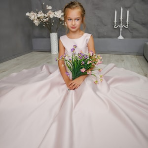 Pu&ograve; includere: Una bambina con un vestito di raso rosa tiene un mazzo di fiori viola e rosa. &Egrave; seduta sul pavimento con le gambe incrociate e guarda la telecamera.