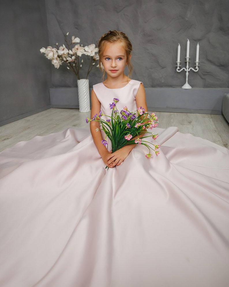 Pu&ograve; includere: Una bambina con un vestito di raso rosa tiene un mazzo di fiori viola e rosa. &Egrave; seduta sul pavimento con le gambe incrociate e guarda la telecamera.