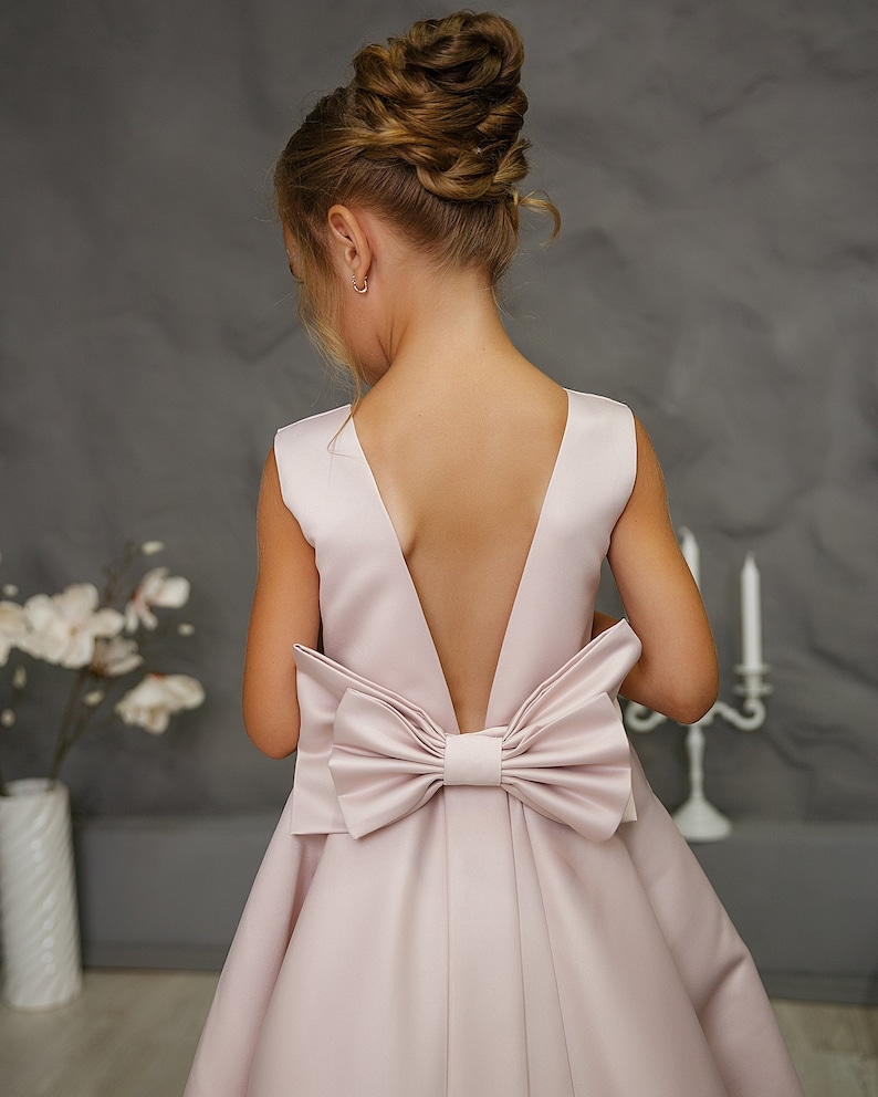Pu&ograve; includere: Un abito di raso rosa chiaro con un grande fiocco sulla schiena. L'abito ha una schiena a V profonda.