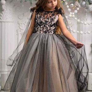 May include: A young girl wearing a black and gray tulle dress with floral embroidery. The dress has a cape and is worn with a silver tiara. The girl is standing in front of a white fireplace decorated with a garland of greenery and silver ornaments. The text "PEARL DRESS BOUTIQUE" is visible at the bottom of the image.