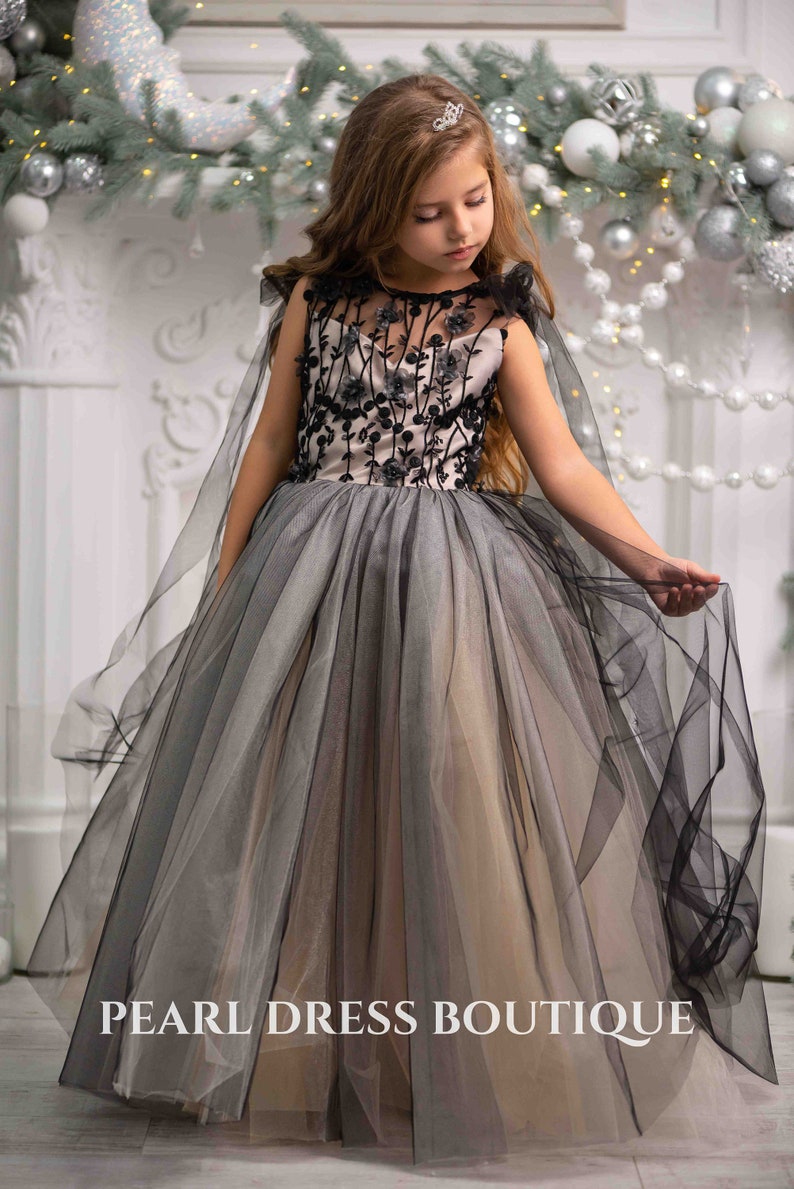 May include: A young girl wearing a black and gray tulle dress with floral embroidery. The dress has a cape and is worn with a silver tiara. The girl is standing in front of a white fireplace decorated with a garland of greenery and silver ornaments. The text "PEARL DRESS BOUTIQUE" is visible at the bottom of the image.