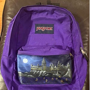 May include: Purple Jansport backpack with a front pocket featuring a painted night scene of a castle on a cliff. The castle is lit up with yellow lights, and the night sky has a full moon. The Jansport logo is on the top.