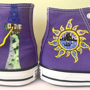 May include: A pair of purple high-top sneakers with hand-painted designs. One shoe features a tower with a yellow braid, while the other has a yellow sun with a castle inside.