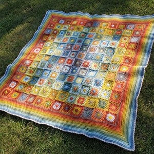 Op de afbeelding: Een kleurrijk gehaakt deken met een regenboogrand. Het deken is gemaakt van vierkanten in verschillende kleuren, waaronder blauw, geel, oranje en rood.