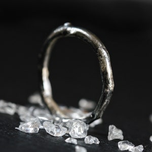 Op de afbeelding: Een zilveren ring met een getextureerde, organisch gevormde band. De ring ligt op een zwart oppervlak met kleine, heldere kristallen die eromheen zijn verspreid.