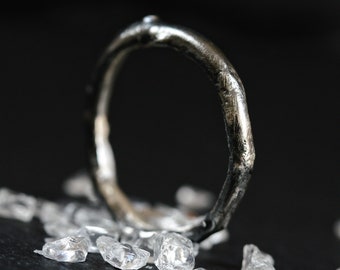 May include: A silver ring with a textured, organic-shaped band. The ring is lying on a black surface with small, clear crystals scattered around it.