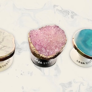 Könnte beinhalten: Drei dekorative Handyhalterungen mit einzigartigen Steindesigns. Einer hat ein weißes Marmormuster, ein anderer einen rosa Kristallcluster und der dritte einen blau-grünen Stein. Jeder hat eine goldfarbene Umrandung und eine weiße Basis mit dem Text "LASA STONES".