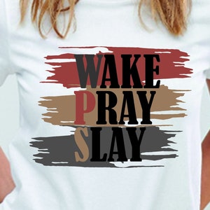 May include: White t-shirt with a graphic print of the words "WAKE PRAY SLAY" in black text on a red, brown, and grey background.