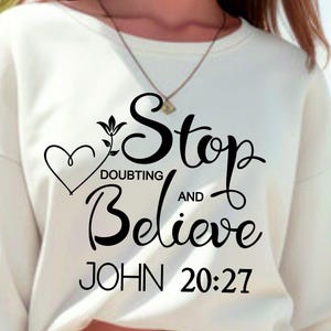 May include: Cream-colored sweatshirt with the words "Stop Doubting and Believe" in a black script font, along with the Bible verse John 20:27. A heart and floral design are incorporated into the text. The sweatshirt is a casual style.