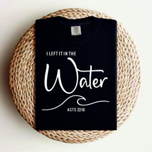 May include: Black t-shirt with white text reading "I LEFT IT IN THE Water", a wave graphic, and "ACTS 22:16". The shirt is folded on a round, woven, beige placemat.