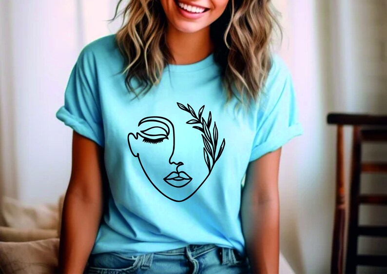 May include: A light blue t-shirt featuring a black line drawing of a face with leaves growing from the eye. The face is closed-eyed and has a simple, minimalist design.
