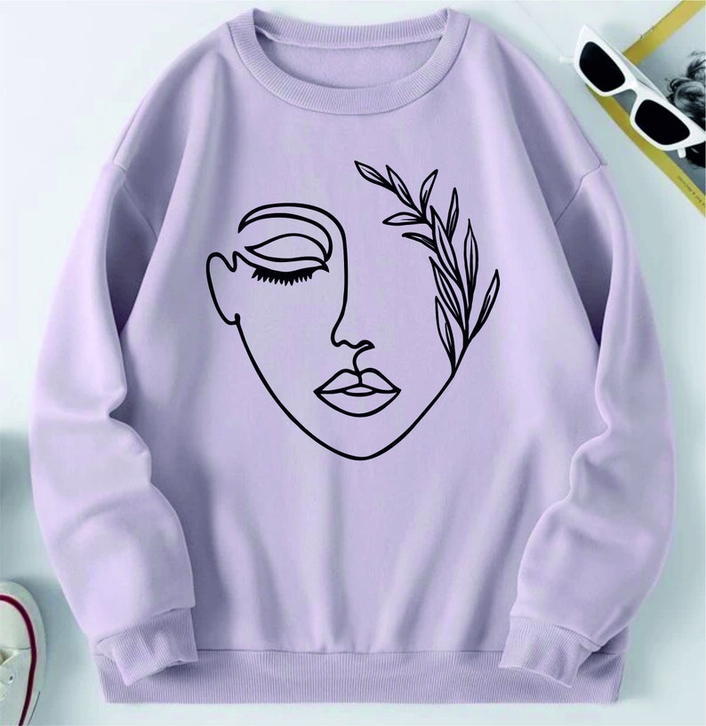 May include: A lavender sweatshirt with a black line drawing of a face with a leafy branch over one eye.
