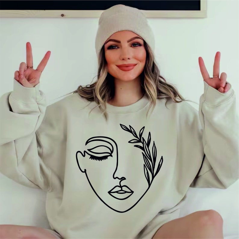 May include: A beige sweatshirt with a black line drawing of a face with a leafy branch growing from the eye. The face has closed eyes and a slightly open mouth.