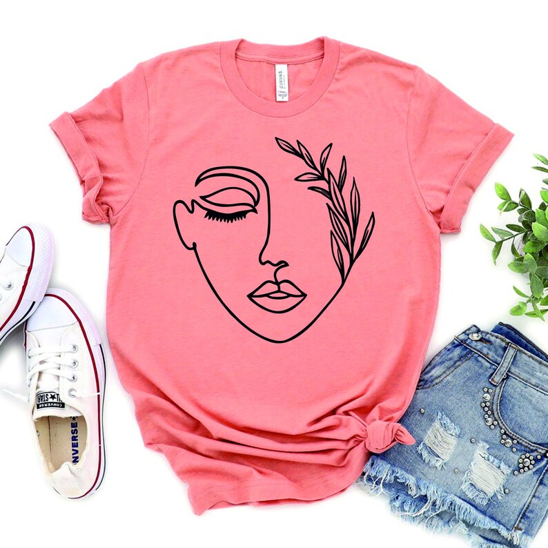 May include: A coral pink t-shirt with a black line drawing of a woman's face with leaves on one side.