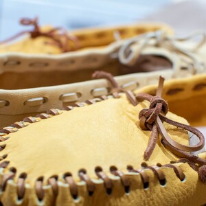 May include: Close-up of two pairs of leather moccasins with brown leather laces. The moccasins are a light tan color and have a decorative stitching detail around the edges.
