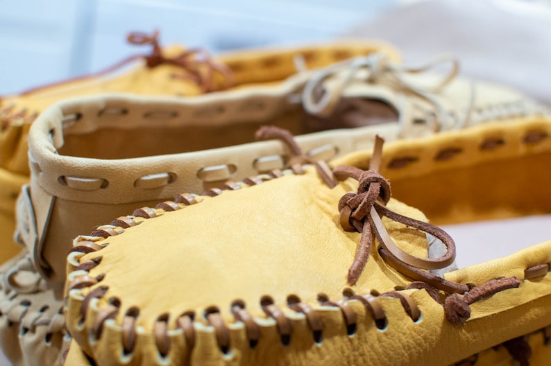 May include: Close-up of two pairs of leather moccasins with brown leather laces. The moccasins are a light tan color and have a decorative stitching detail around the edges.