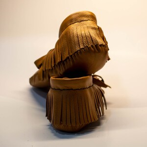 May include: A pair of brown leather moccasin-style baby shoes with fringe detailing. The shoes are made of soft leather and have a classic design. The fringe adds a decorative element to the footwear.
