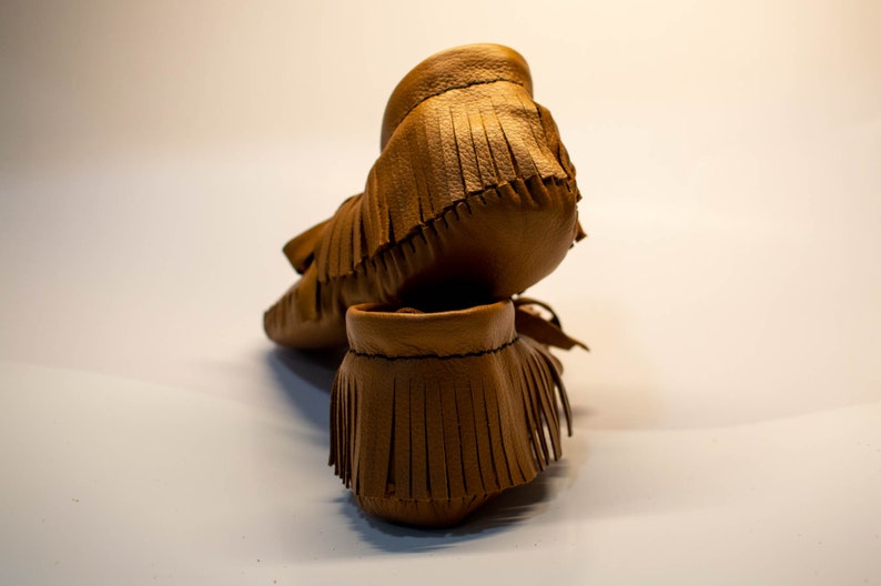 May include: A pair of brown leather moccasin-style baby shoes with fringe detailing. The shoes are made of soft leather and have a classic design. The fringe adds a decorative element to the footwear.