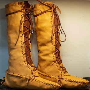 May include: A pair of tall, tan leather boots with brown laces. The boots have a moccasin-style toe and are laced up the front.