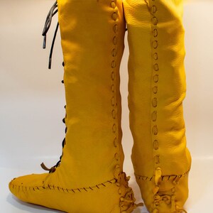 May include: A pair of yellow leather knee-high boots with laces up the front and back. The boots have a moccasin-style toe and are stitched with a decorative pattern.