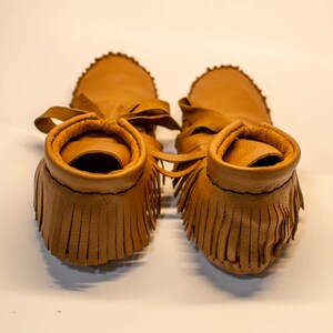 May include: A pair of tan leather moccasin-style baby shoes. The shoes have a fringed vamp, a soft sole, and a tie closure. The edges of the shoes are stitched with black thread. The shoes are designed for infants.