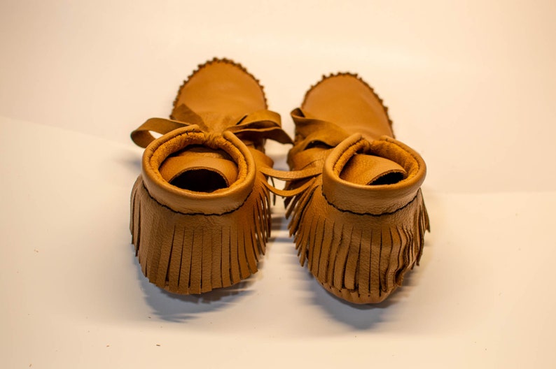 May include: A pair of tan leather moccasin-style baby shoes. The shoes have a fringed vamp, a soft sole, and a tie closure. The edges of the shoes are stitched with black thread. The shoes are designed for infants.