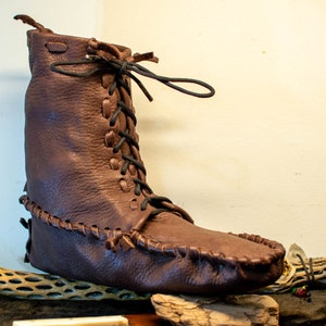 Puede incluir: Una bota mocas&iacute;n de cuero marr&oacute;n con cierre de cordones. La bota tiene una puntada decorativa alrededor de la suela y una punta puntiaguda.
