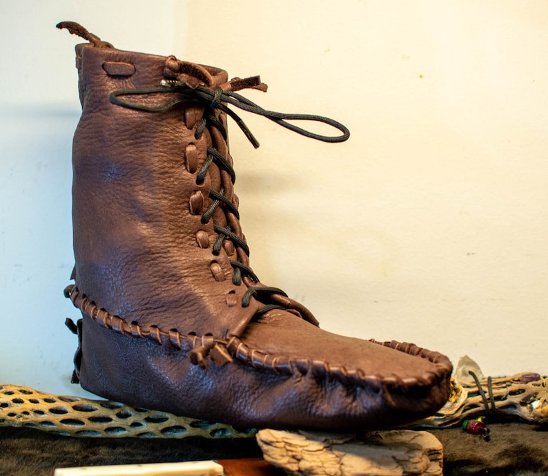Puede incluir: Una bota mocas&iacute;n de cuero marr&oacute;n con cierre de cordones. La bota tiene una puntada decorativa alrededor de la suela y una punta puntiaguda.