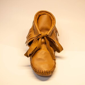 May include: A single tan leather moccasin with a fringed upper and a tied bow. The shoe has a rounded toe and is stitched along the sole. The moccasin is designed for infants or toddlers.
