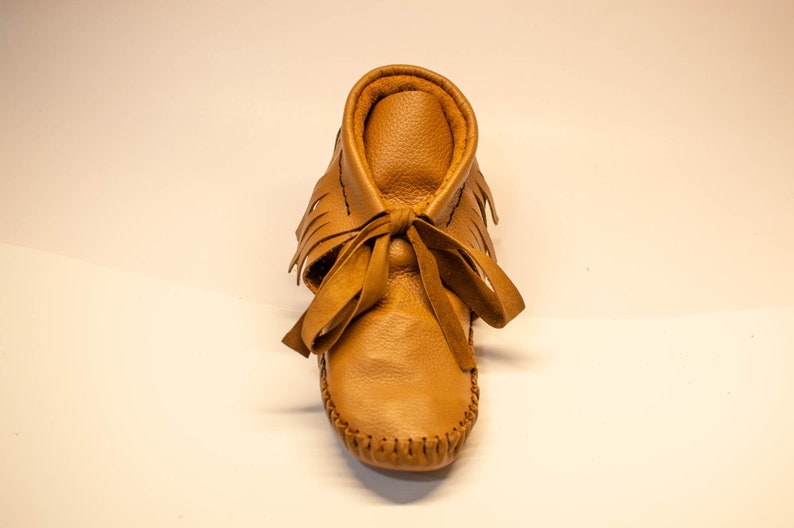 May include: A single tan leather moccasin with a fringed upper and a tied bow. The shoe has a rounded toe and is stitched along the sole. The moccasin is designed for infants or toddlers.