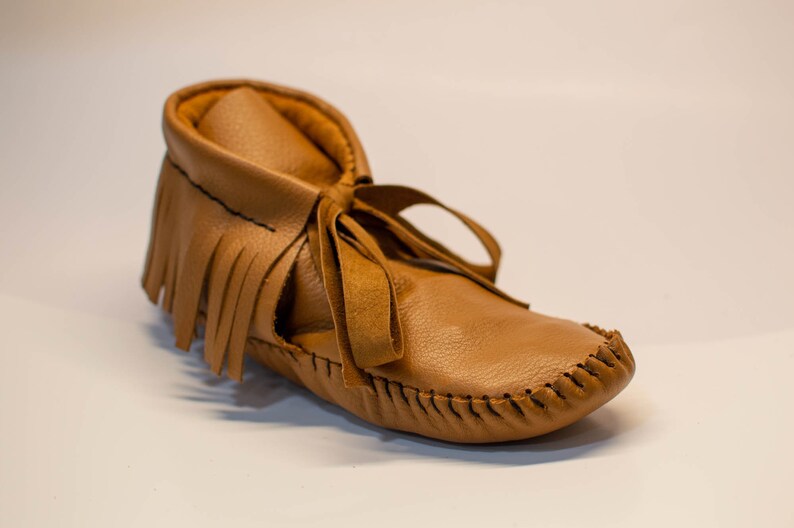 May include: A tan leather moccasin-style baby shoe with a fringed upper and a tie closure. The shoe features a stitched sole and a soft, flexible design, suitable for infants and toddlers. The footwear is designed for comfort and style.