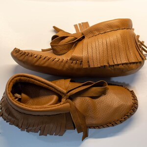 May include: A pair of brown leather moccasin baby shoes. The shoes have a soft, textured appearance with fringe detailing around the ankle and a bow on the top. The shoes are designed with a classic, handcrafted style.