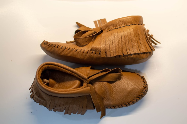 May include: A pair of brown leather moccasin baby shoes. The shoes have a soft, textured appearance with fringe detailing around the ankle and a bow on the top. The shoes are designed with a classic, handcrafted style.
